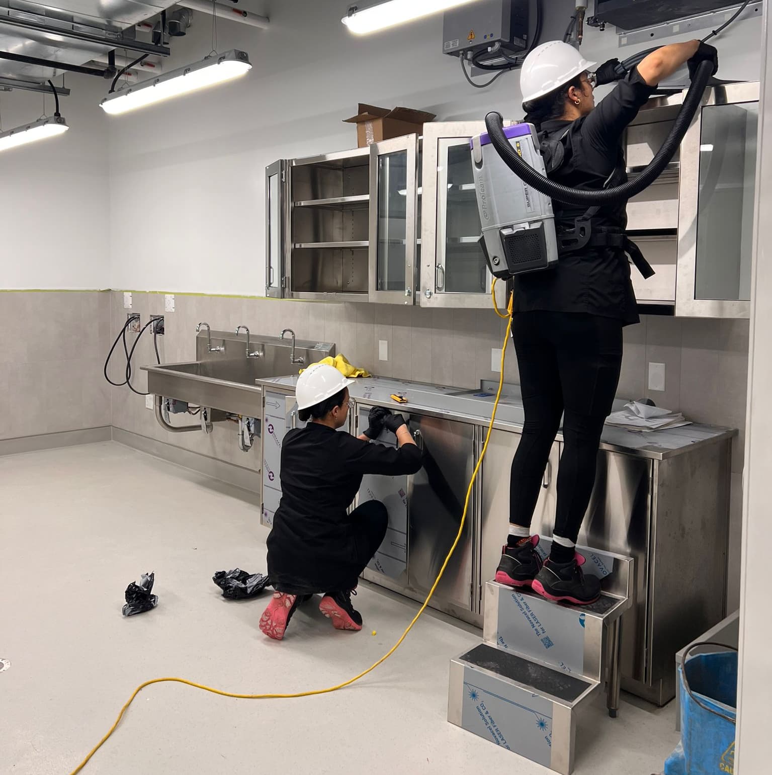 Two Flores Clean team members in uniform and hard hats deep-cleaning a stainless-steel commercial kitchen, one vacuuming the ceiling vents and the other wiping down cabinetry
