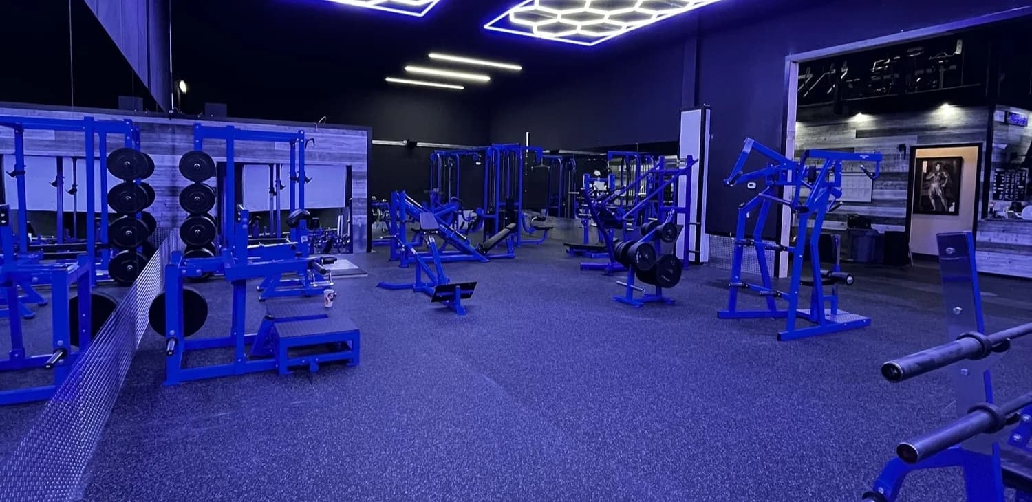 A blue-lit strength-training gym with rows of plate-loaded machines and free weights