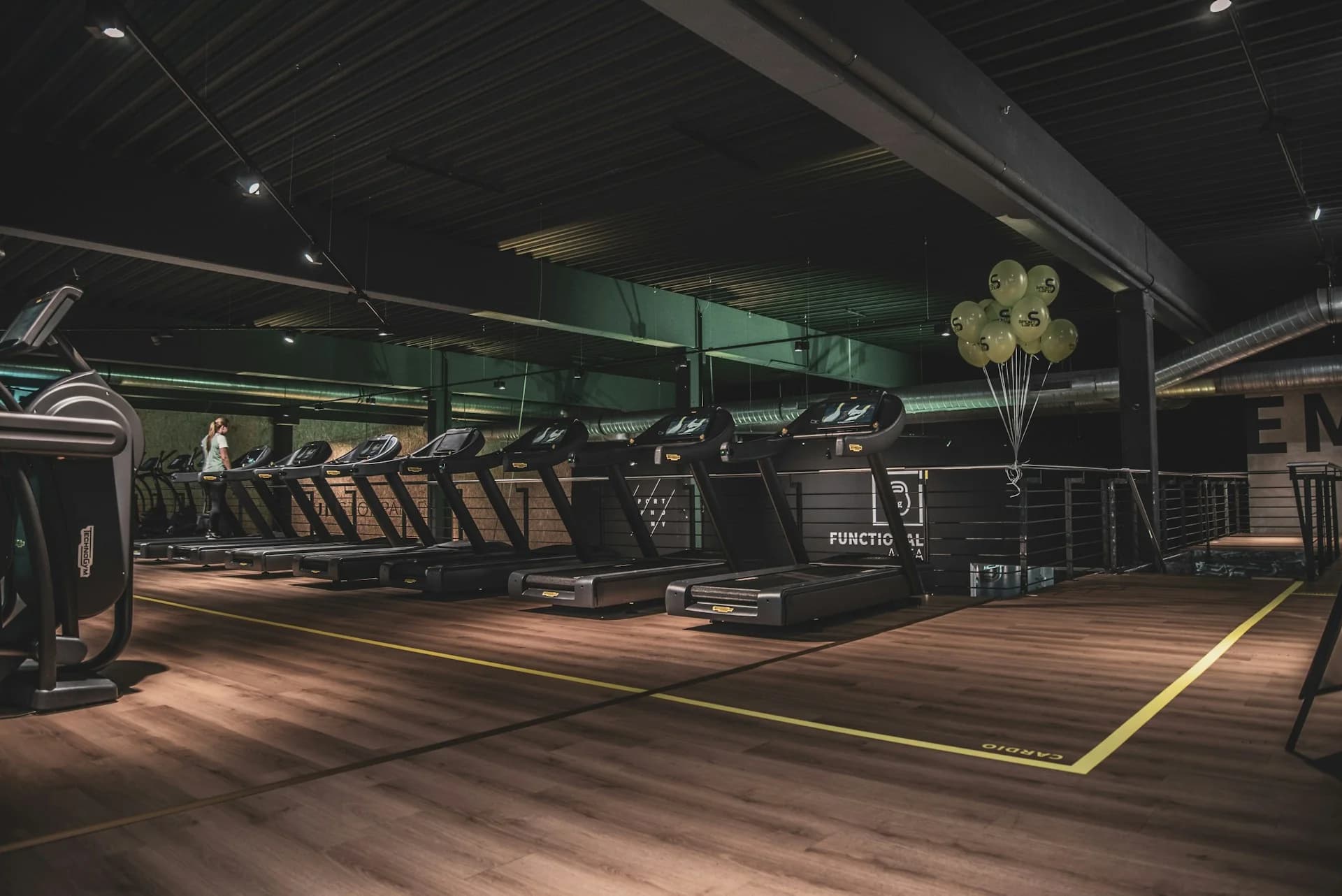 A dark, modern commercial gym with a long row of treadmills and cardio machines under moody lighting