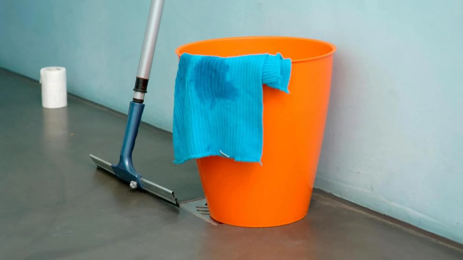An orange cleaning bucket with a microfiber cloth and a floor squeegee against a wall
