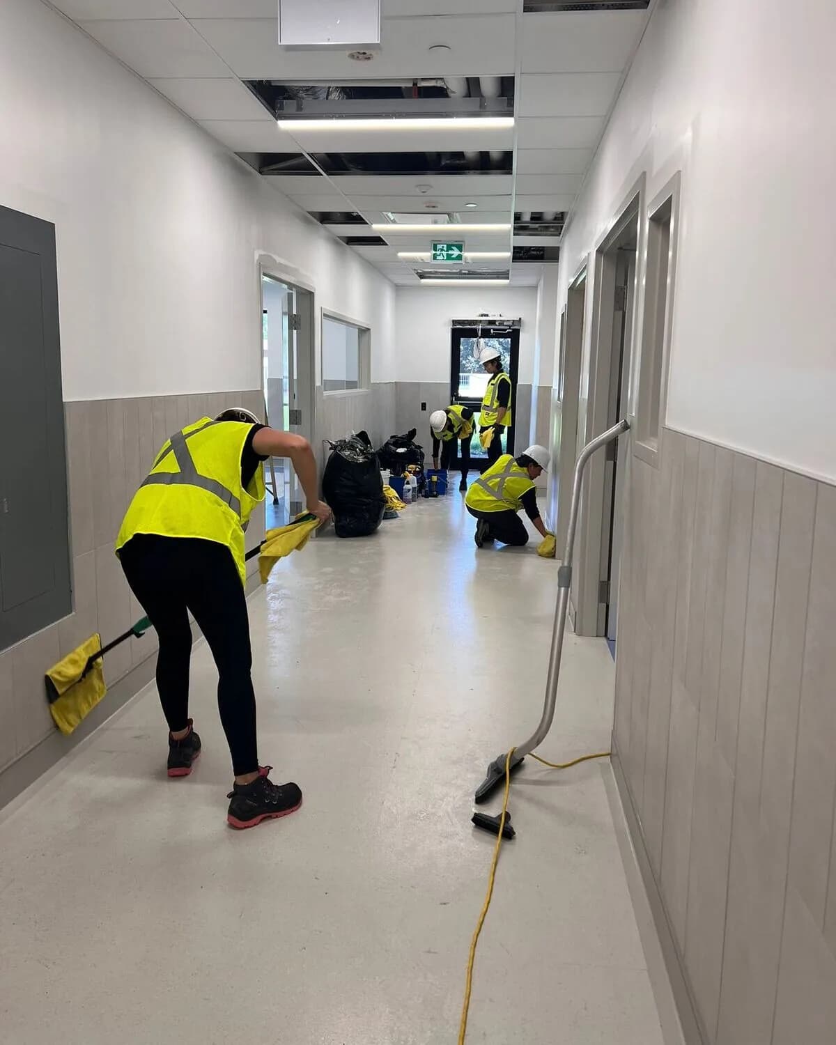 A cleaning crew in hi-vis vests and hard hats mopping and vacuuming a corridor during a post-construction cleanup