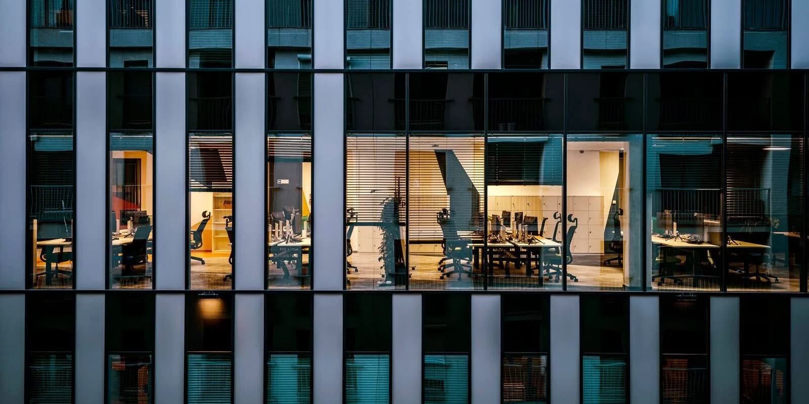 Lit open-plan offices seen through the clean glass facade of a commercial building at dusk