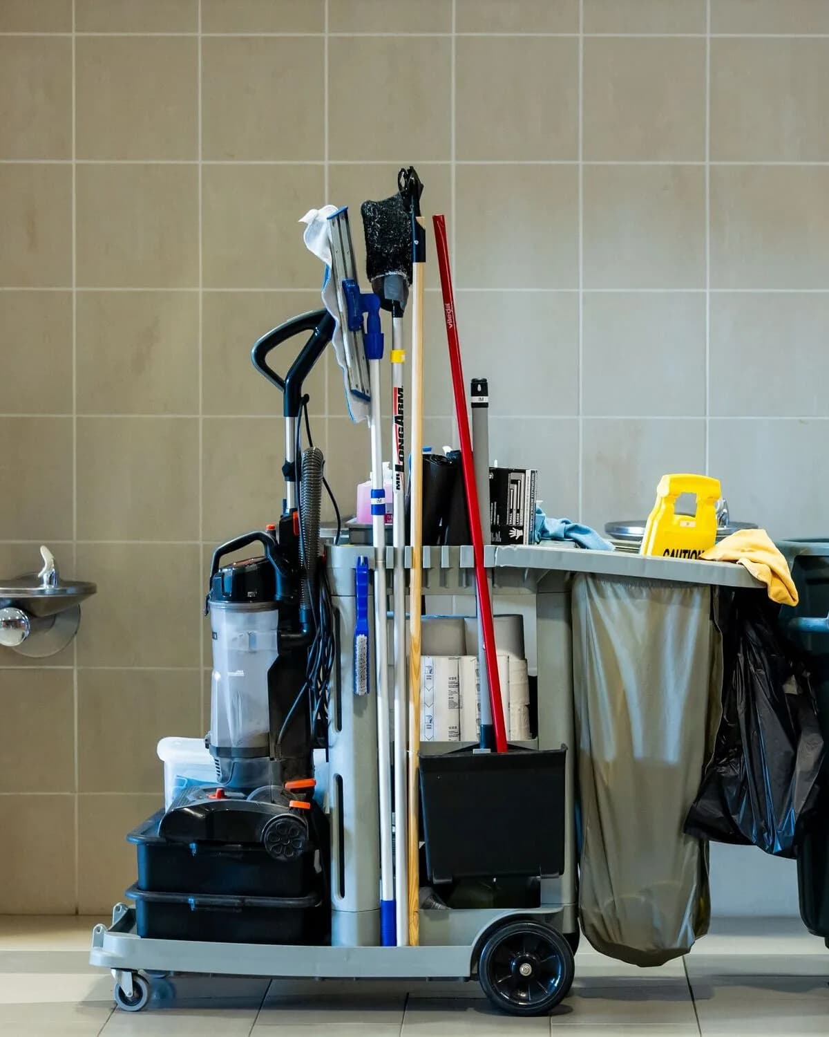 A fully stocked janitorial cart with mop, broom, vacuum, and cleaning supplies beside a tiled wall