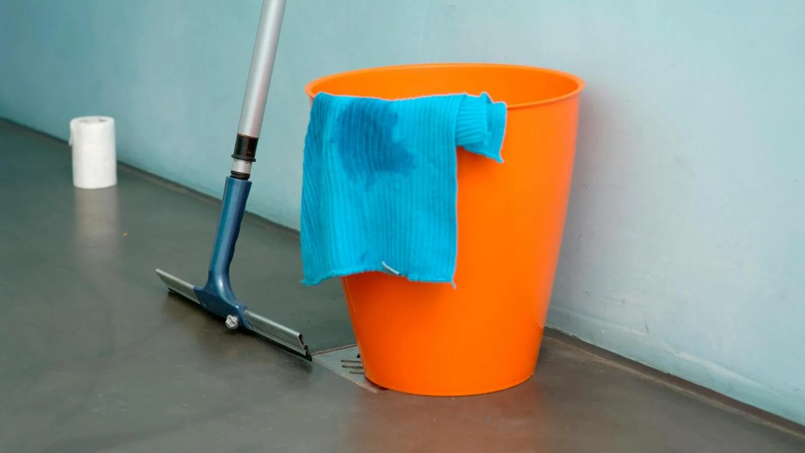 Floor squeegee and orange mop bucket with cleaning cloth against a wall